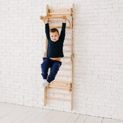 Child climbing a wooden wall climber against a white brick wall.